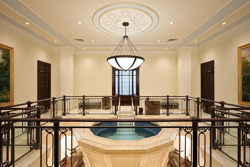 Interior photo of the Baptistry at the Latter-Day Saints temple. A large limestone pool centers the room with bronze railing surrounding.